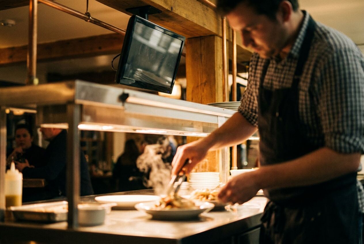 A cook plates a dish under heat lamps at a timber-framed kitchen pass.